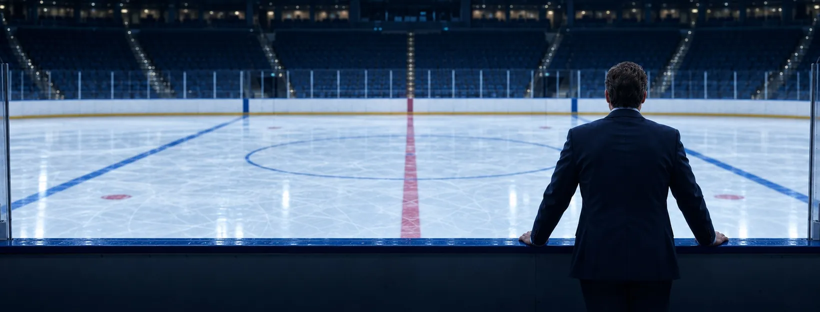 A coach seen from behind at the boards, looking across the ice with both blue lines in view.